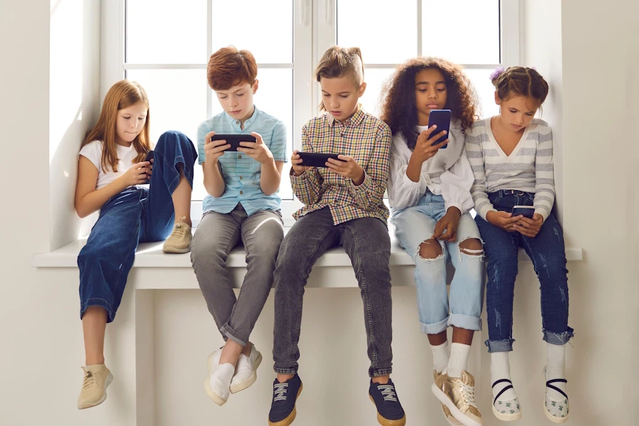 Five children sitting on a windowsill, each looking at their own mobile phone.