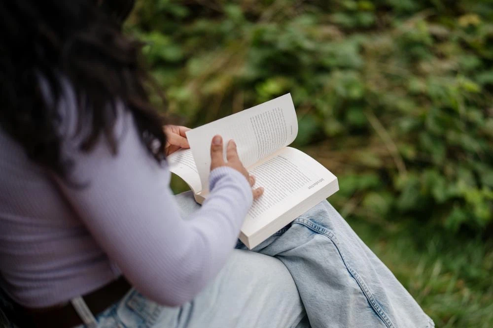 The camera looks over the shoulder of a woman sitting on a bench and reading a book.